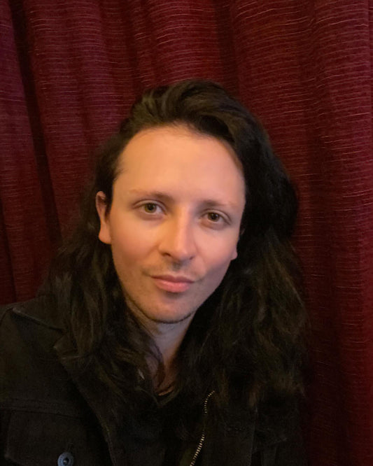 Old Town Magick founder, Marcus, with long, dark hair and a black jacket stands against a maroon curtain, evoking a gothic, mysterious mood.