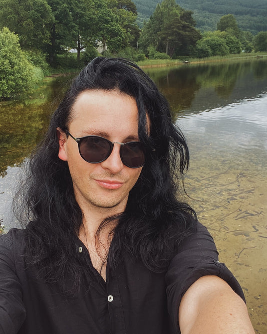 A person with long black hair and sunglasses poses for a selfie near a lake, reflecting the blog's gothic aesthetic.
