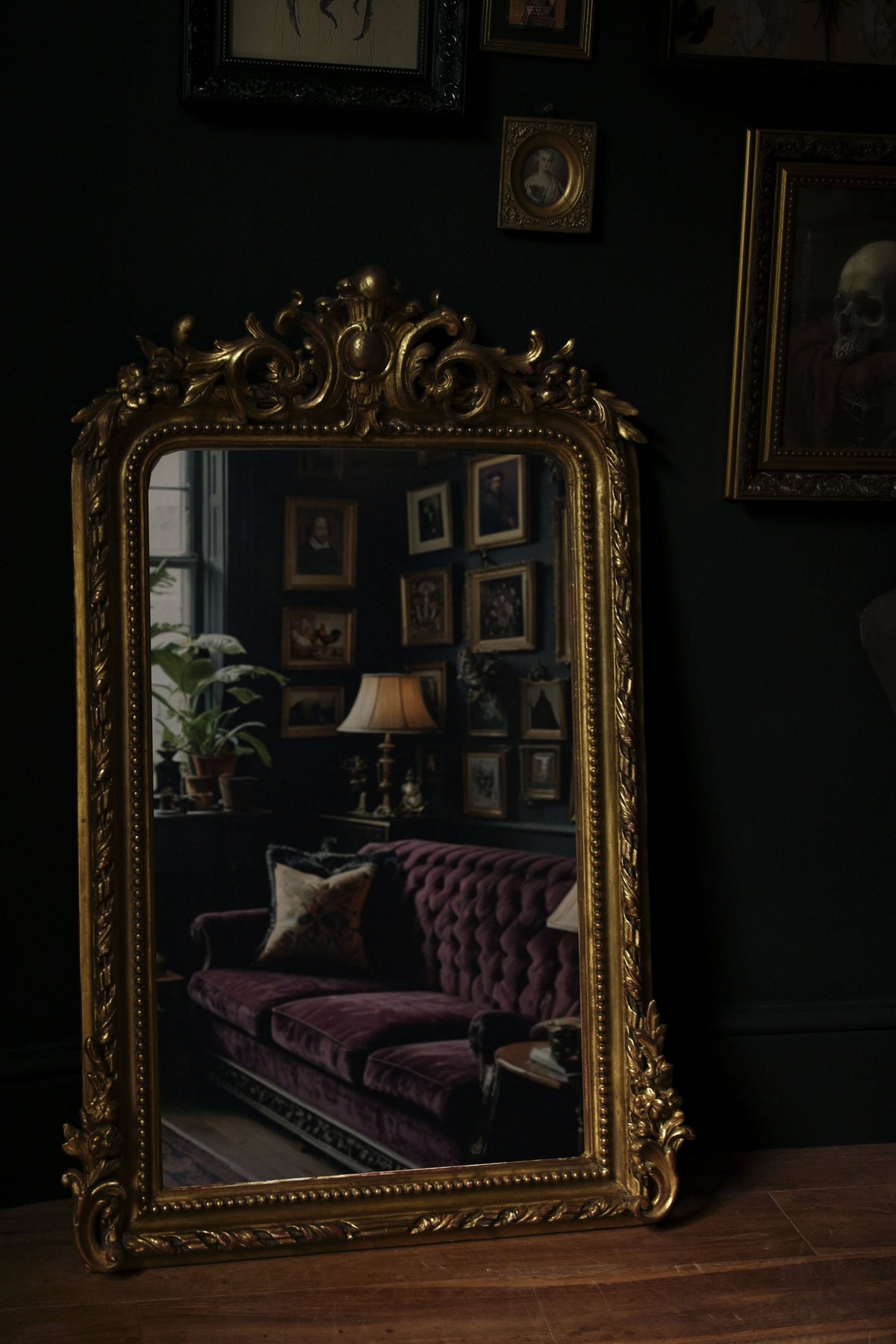 Ornate gold mirror reflects a dimly lit room with a purple velvet couch and dark walls adorned with framed pictures.
