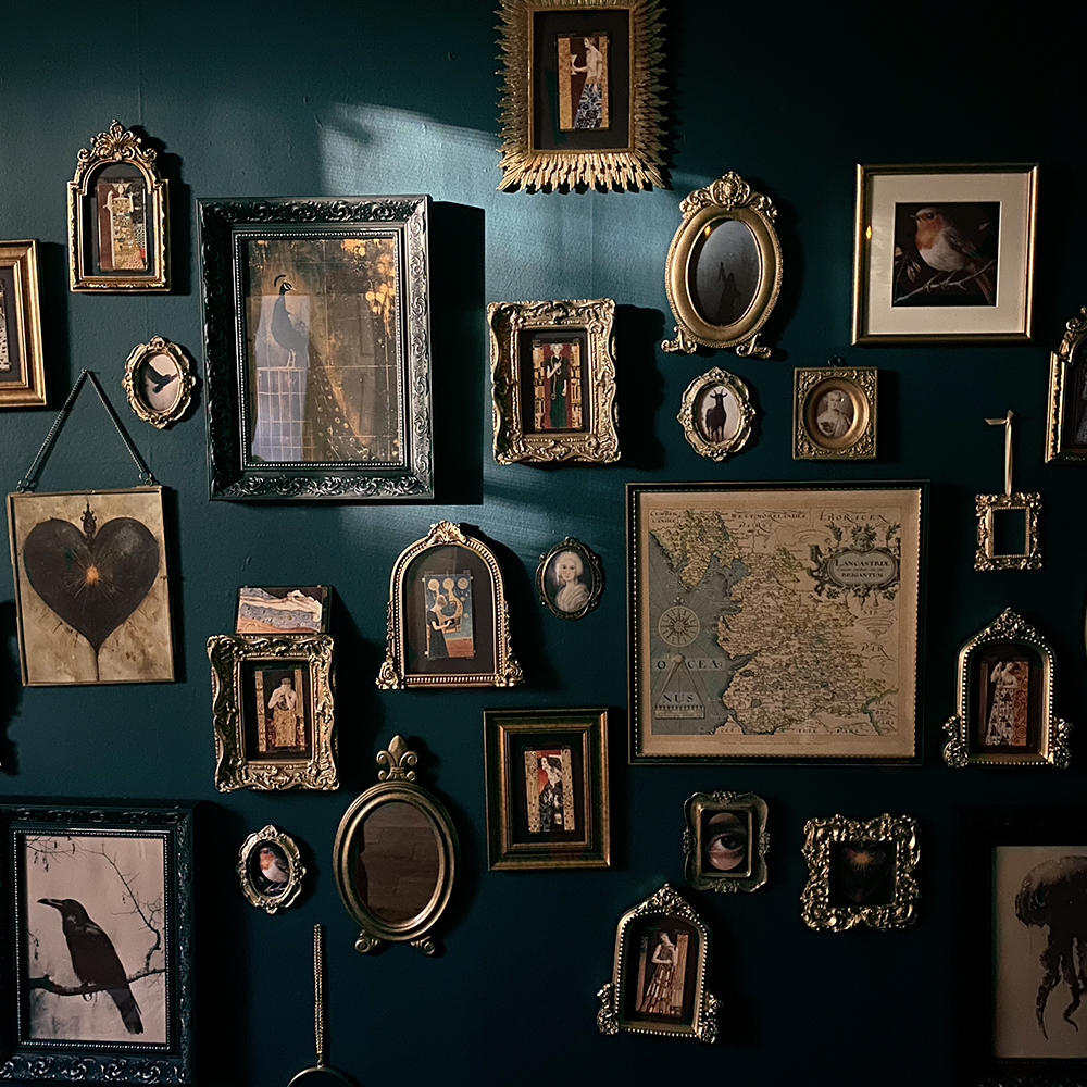 A dark teal wall is covered in a collection of ornate, gold-toned framed pictures and mirrors of varying sizes. A shaft of light illuminates the top of the wall.