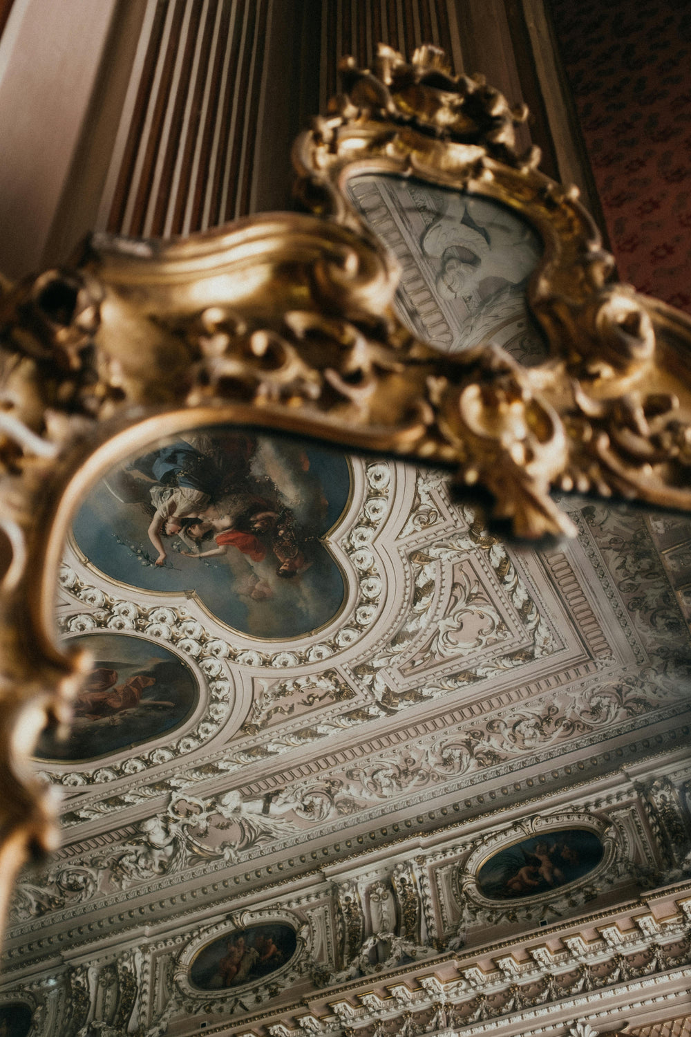 Ornate gold mirror reflecting an elaborate ceiling with paintings, carvings, and molding. Ceiling features blue and white with pink accents.
