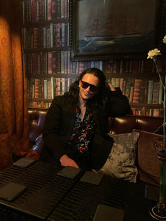 A person with long dark hair and sunglasses lounges on a leather sofa in a dimly lit room with a bookshelf backdrop.