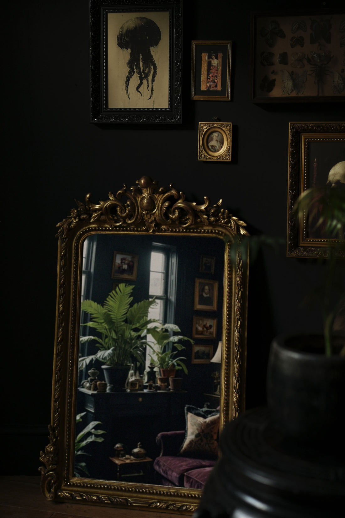 Ornate gold mirror reflects a dark room with plants, desk, and chair, against a dark wall adorned with framed art.

