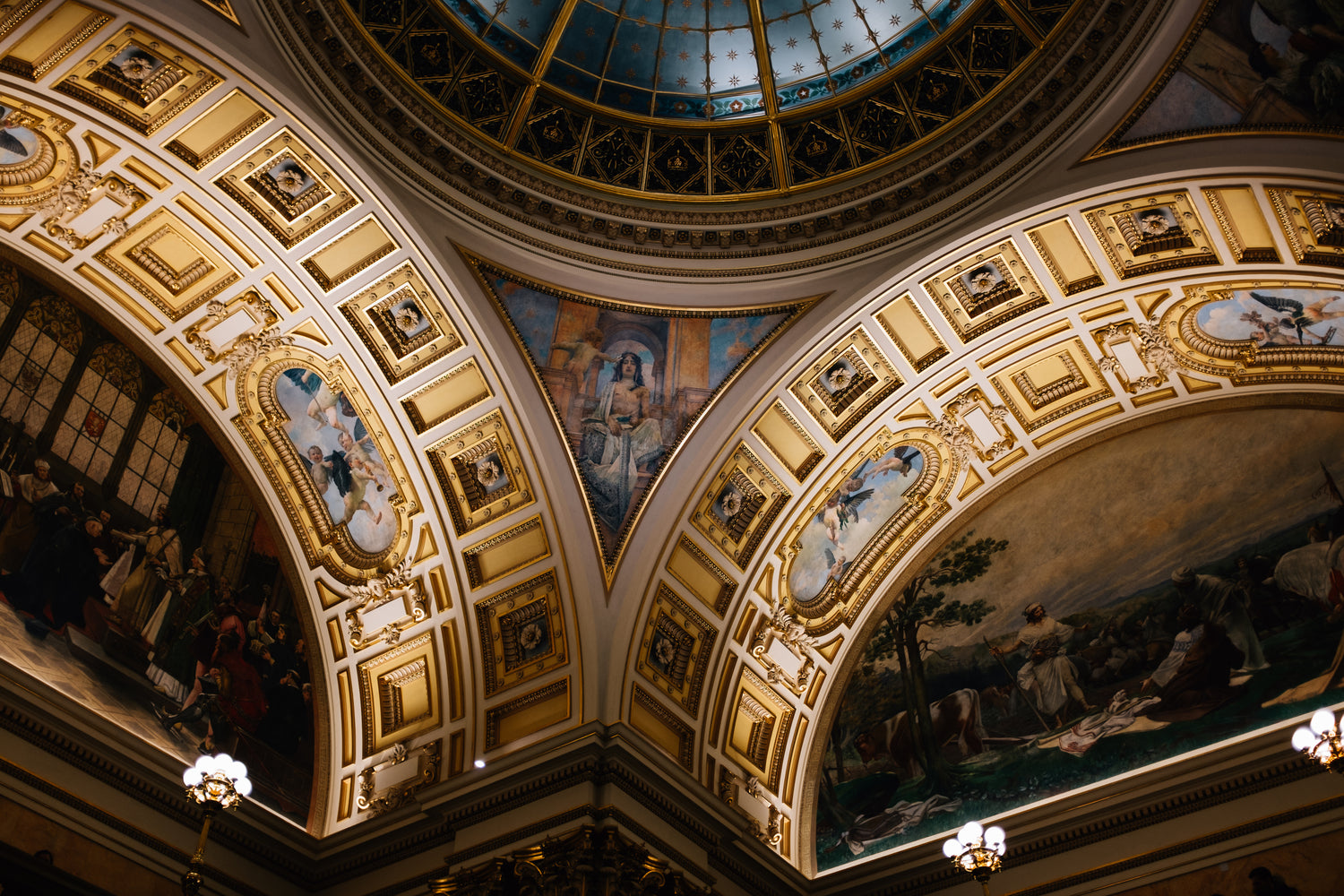 Ornate ceiling featuring murals and gold-trimmed architectural details, illuminated with warm light. A blue glass dome sits at the apex.