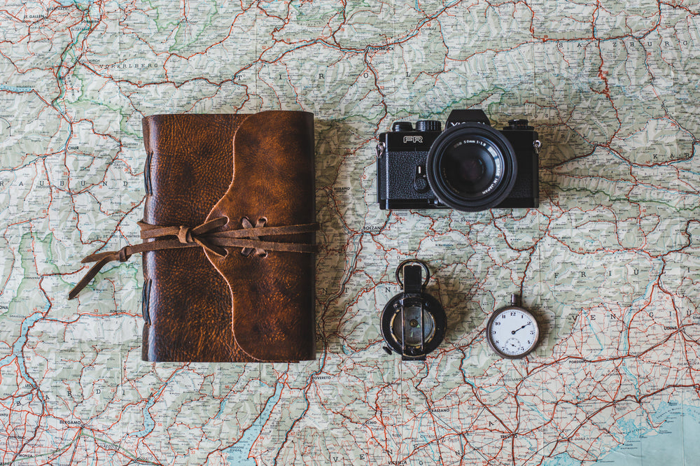 Overhead shot of a map with a brown leather-bound journal, black camera, compass, and pocket watch arranged on top.