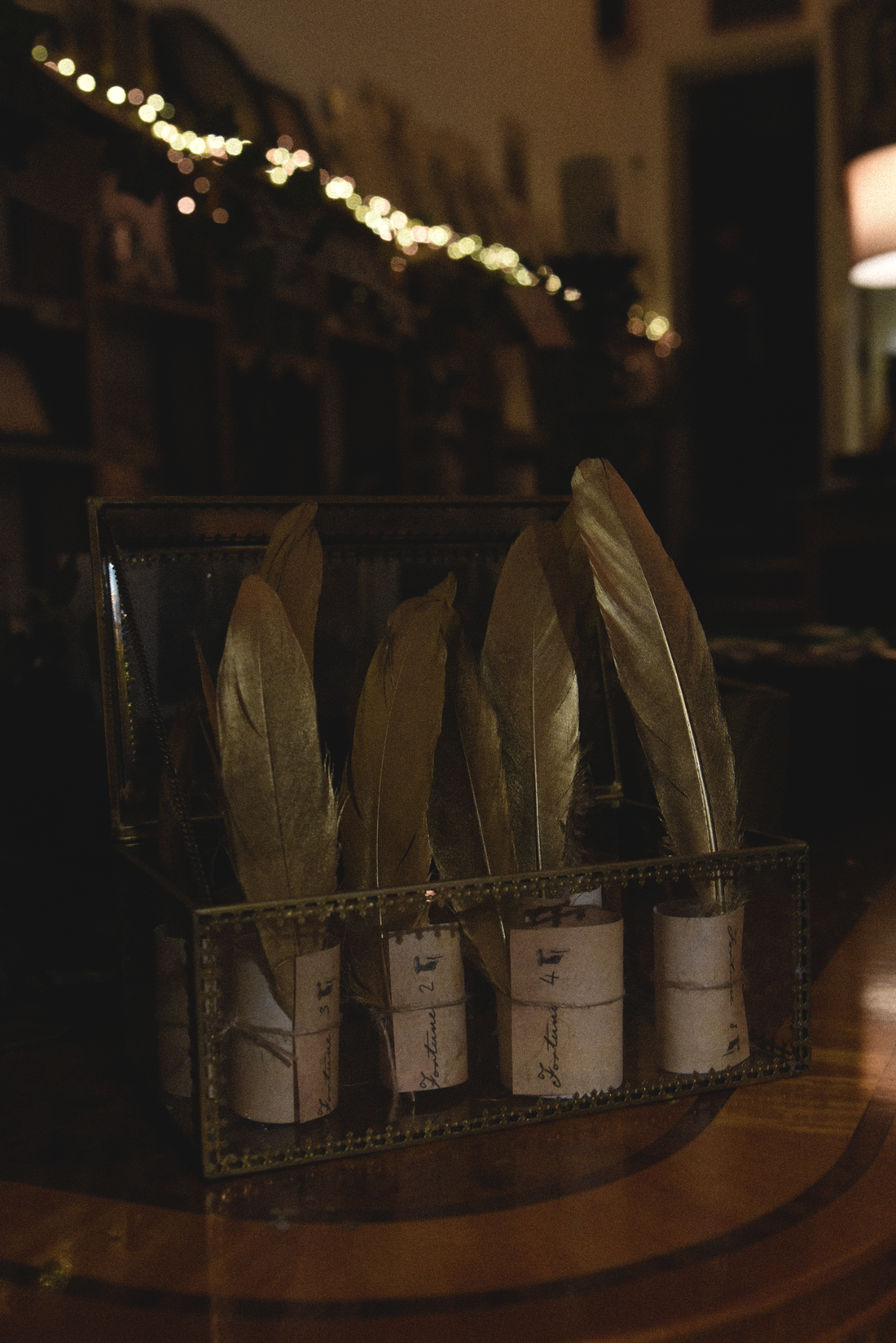 A glass box contains four gold feathers, each secured with a wrapped "fortune" label; string lights blur in the dark background.