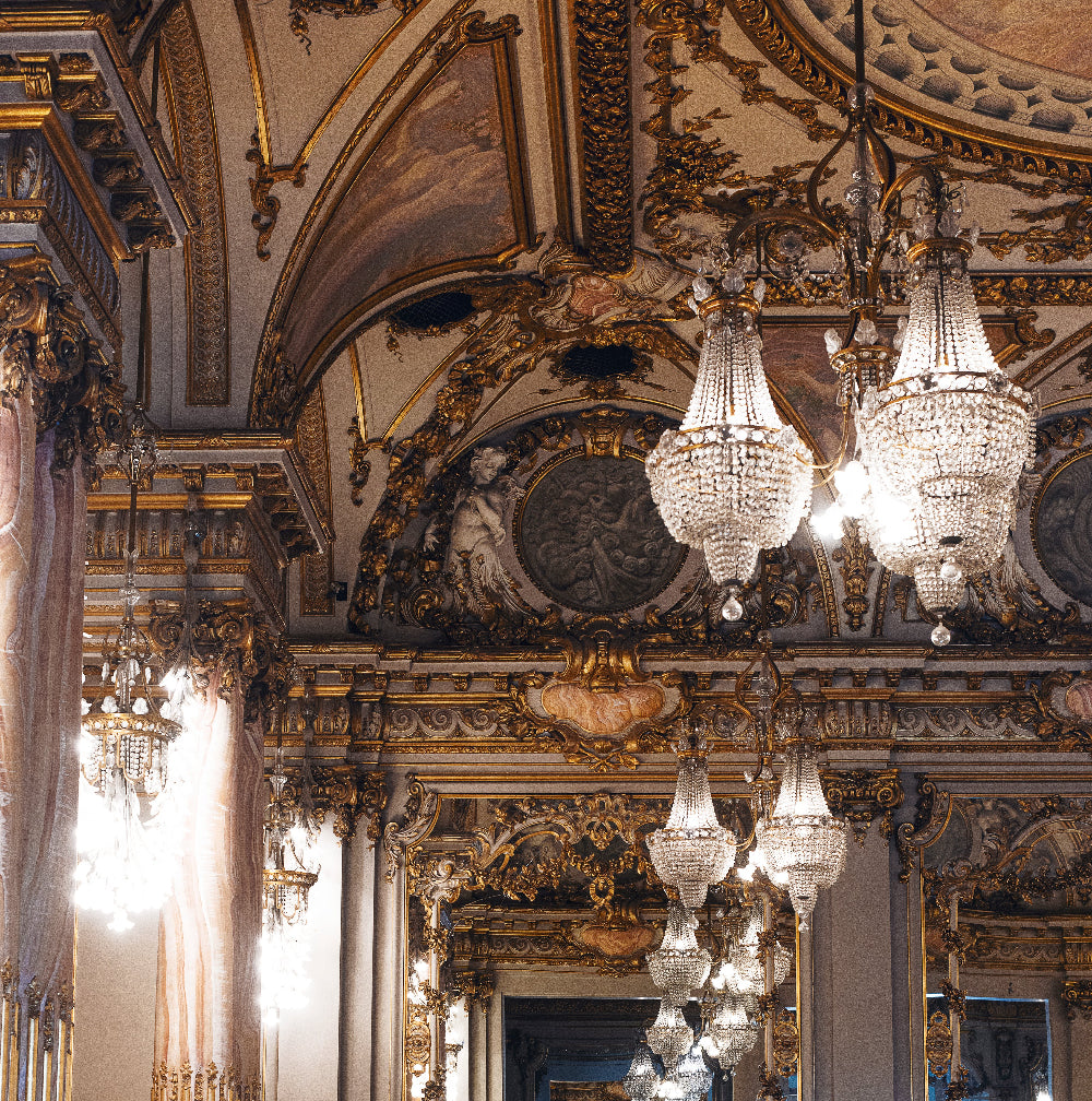 Opulent room interior with multiple crystal chandeliers, gilded baroque detailing, and ornate ceiling art. Warm, elegant lighting accentuates the classical architecture.